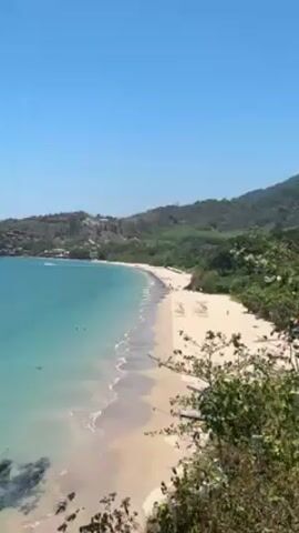 The video shows a panoramic view of a tropical beach on a sunny day. The sky is clear and blue. The water is turquoise and meets a sandy beach with gentle waves. Some people are visible on the beach and in the water. A forested hill forms the backdrop of the beach. In the distance, there is a rocky headland with a building atop it, surrounded by several boats. A tree with light-colored branches is in the foreground on the right side of the screen.