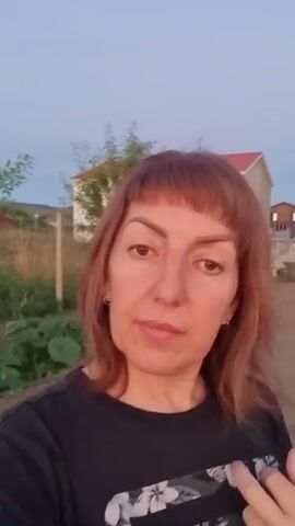 A medium shot video begins with a woman looking at the camera outdoors. She has shoulder-length reddish-brown hair with bangs. She is wearing a black t-shirt with a white floral pattern. Her left hand is raised, and her index finger is pointing at the t-shirt. Behind her, there is vegetation, a wooden fence post, and a house. The sky is visible in the background. The video continues with the woman slowly moving her head from left to right and her finger moving around on her shirt. The environment appears to be a rural or garden area, possibly at dusk.