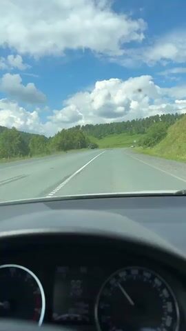 This is a first-person perspective video, likely from a dashcam, showing a drive through a rural landscape under a bright, cloudy sky. The scene is a paved road with a double white line marking the center, suggesting a two-lane highway. The road curves gently to the right, leading towards rolling green hills dotted with trees. In the distance, the landscape appears to be a mix of grassy fields and dense forests, with a few small trees scattered on the hillsides. The sky is a vibrant blue, filled with large, fluffy white clouds.

On the left side of the road, there are trees and some greenery. A dark-colored SUV is parked on the shoulder of the road. On the right side, there are more trees and roadside vegetation, as well as a road sign that appears to have symbols indicating a hazard or a specific road condition. Further down the road, a few more vehicles are visible in the distance, also traveling in the same direction.

The interior of the vehicle is visible in the foreground, showing the dashboard. A speedometer is visible on the right, with the needle indicating a speed around 90. A digital display in the center shows "90" prominently, likely representing the current speed in kilometers per hour, along with other unreadable data.

The overall impression is a scenic drive on a sunny day in a rural area, possibly in Russia given the location context of Бердагулово. The weather appears to be pleasant with a mix of sun and clouds. There are no people visible in the frame, only vehicles and the landscape. The mood conveyed is one of peaceful travel and enjoying the scenery.