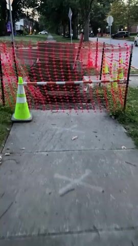 The video shows a construction site in a residential area. A trench has been dug in the sidewalk and yard, with a large hole filled with murky water in the center. Red plastic fencing and orange traffic cones surround the area, indicating a hazardous zone. A wooden support structure has been placed over the trench, and a ladder leans against it. The street and houses are visible in the background, suggesting an urban or suburban setting. The lighting indicates it is either early morning or late evening.