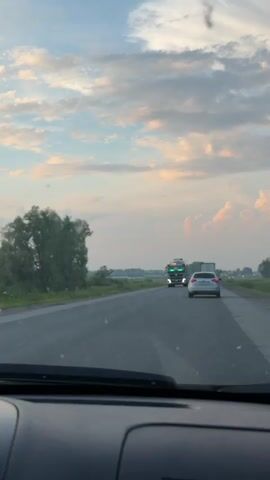 A video shows a view from inside a vehicle driving on a highway. The road has at least two lanes and is lined with trees and fields on the left side. Several vehicles are on the road, including a cement mixer truck with green lights above its windshield, a white car, and other cars in the distance. The sky is partly cloudy with a mix of blue and white and some pale orange hues, suggesting it might be dawn or dusk. The camera appears to be mounted inside the vehicle, looking forward through the windshield.