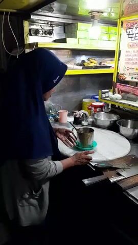 A person wearing a dark blue hijab and a grey hoodie is standing behind a food cart, preparing a drink in a metal cup. The cart is illuminated by overhead lights. In the background, there is a menu board with various food items and prices listed. A large griddle is visible in the foreground, with a circular food item being cooked on it. Various bowls, containers, and utensils are arranged on the counter. The environment appears to be an outdoor setting at night, with a dark street visible beyond the cart.