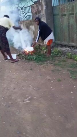 Two individuals are standing on a dirt path next to a small, controlled fire. One individual, wearing a yellow top and a dark skirt with colorful patterns, bends over to tend to the fire. The other individual, wearing a dark hoodie and orange shorts, is also near the fire, holding a light-colored object over it. The fire is producing a significant amount of white smoke. In the background, there is a wall and some green foliage.