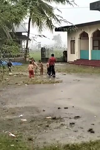 The scene shows several children playing outside in the rain near a building that appears to be a mosque, based on its architecture featuring arches. A man on a bicycle is present amidst the children, who are scattered around a grassy, somewhat muddy area with litter. Some of the children are shirtless.  Palm trees are visible in the background. The overall atmosphere suggests a rainy day in a rural or semi-rural location.