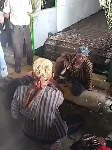 The video captures an outdoor or semi-open gathering in Sragen, Indonesia, during daylight hours. In the foreground, two individuals are seated on the ground. One person, with light blonde hair and a brown headband, wears a striped tunic. Next to them, another individual with dark hair and a head covering, also in a striped top, holds a small stick or pipe. Both appear focused, suggesting they are participating in a cultural performance or ritual.

In the background, several men stand observing. One man wears a light shirt and light blue pants, while another in a light shirt, brown pants, and a cap watches intently. Part of a person or a prop, with dark hair and red fabric, is visible lying on the ground in the bottom right corner. The setting includes a white wall with green accents, and a raised wooden or metal grated platform behind the seated individuals. Decorative palm fronds or similar foliage hang overhead. The ground beneath them is concrete, scattered with small white debris or petals. There is no visible text. The overall atmosphere appears calm and observant.