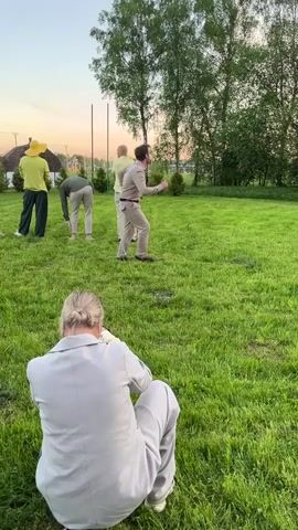 The video shows a person in a khaki safari-style outfit dancing on a grassy lawn. Other people are standing or sitting around watching. One person in a gray suit is crouched down in the foreground, possibly taking a photo. There are trees in the background and a house visible in the distance. The sky is dusky.