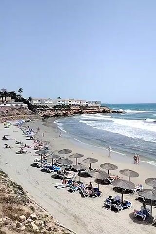 The video shows a crowded beach with many people sunbathing on lounge chairs under thatched umbrellas. The ocean has moderate waves, and a few people are walking along the shoreline. Buildings are visible on a cliff overlooking the beach. The sky is clear and blue. The video is shot from a high angle, looking down at the beach and the ocean.