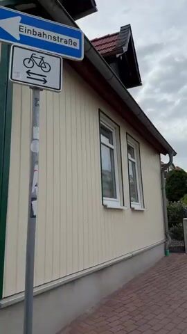 The video shows a slow panning shot of a residential area. The first frame shows a close-up of a window on a tan-colored house with white trim. The second frame pans to the right to reveal a mailbox and the house number "36" next to the window. The third frame continues panning right to show a green garage door and a stone pillar. The final frame shows a streetlamp with a cloudy sky and houses in the background. The overall scene depicts a quiet suburban neighborhood with typical residential structures. There are no people or animals visible in the video.