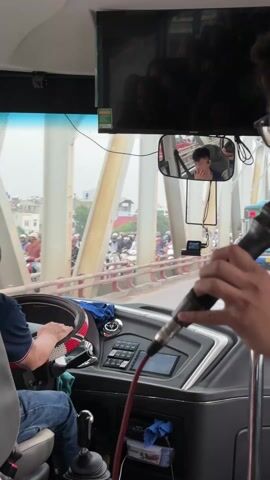 A video shows the view from the front interior of a bus, driving over a bridge. The bus driver is wearing a blue shirt and blue jeans. In the rearview mirror, a young man is visible, seemingly talking. The video is being recorded by someone holding a microphone with a red cable. Another person’s arm gestures to the right of the bus, indicating various vehicles on the road, including a blue bus and a light green car. The bridge has white steel support structures and a railing separating lanes. Traffic consists of cars and motorcycles. A television screen above the driver shows a dark reflection. The bus has a dashboard with various controls and a digital screen. The steering wheel has a red and white cover.