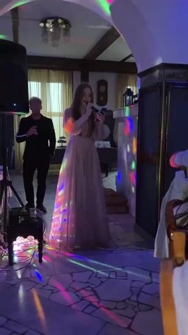In a dimly lit indoor venue with a stone floor, a young woman with long, reddish-brown hair, wearing a long, sparkling light pink dress with puff sleeves, stands center frame, singing into a microphone while looking at a phone in her hand. Colorful lights are projected onto the floor and her dress. Behind her and to the left, a man in a black suit and black sneakers, also holding a microphone, stands facing slightly away from the camera. The setting appears to be a restaurant or event hall, with a chandelier visible on the ceiling, wooden beams, and draped curtains in the background. To the right, there's a fireplace or decorative wall structure. The mood seems festive, possibly a performance or karaoke event. The location context is Zavod Mikhailovskii, Russia.