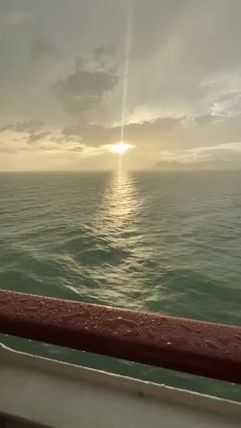 This video captures a serene sunset over the Gulf of Thailand, viewed from the deck of a ferry. The sun, a bright, intense orb, peeks through a sky filled with dramatic, textured clouds, casting a golden, shimmering reflection across the choppy, turquoise water. The rain appears to be starting, with visible streaks of water droplets in the air, adding a subtle yet palpable atmosphere to the scene. In the foreground, a red railing is speckled with raindrops, indicating the onset of the weather change.

The overall mood is one of tranquil beauty, tinged with the slight melancholy often associated with fading daylight and approaching rain. The vast expanse of the ocean, meeting the distant, hazy silhouette of land or islands, evokes a sense of journey and contemplation. The time of day is clearly late afternoon or early evening, as indicated by the low angle of the sun and the warm, golden light. The location context of Don Sak, Thailand, suggests a transition, perhaps as the ferry is departing or arriving at a port, connecting the mainland to one of Thailand's southern islands. The only visible "text" is the timestamp in the video, "00:00," at the beginning.