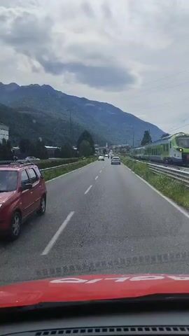 The video shows a view from inside a vehicle driving on a paved road. A train track runs parallel to the road on the right side, and a green and white train is visible on the tracks. Mountains are in the background, and the sky is overcast with clouds. Several cars are driving on the road ahead. The dashboard of the vehicle, painted red, is visible at the bottom of the frame.