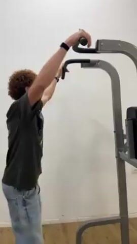A young man with curly brown hair is shown from the side as he performs a pull-up on a pull-up bar. The machine is grey and is in front of a white wall. The man is wearing a dark green shirt and light blue jeans. He has a black watch on his wrist. He pulls himself up and then slowly lowers himself.