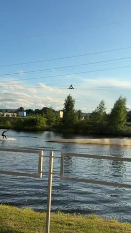 A wakeboarder is being pulled across a body of water by a cable system. The wakeboarder is wearing dark clothing and is in a crouched position. The water is choppy, and there is a spray of water behind the wakeboarder. In the background, there are trees, buildings, and power lines. The sky is blue with some clouds.