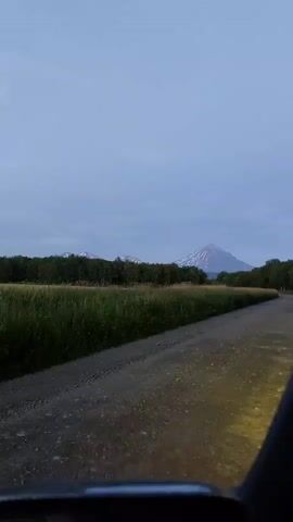 The video shows a dirt road leading towards a conical mountain with snow-capped peaks. A line of trees and tall grass borders the road on the left. The sky is overcast with a pale blue hue. The viewpoint appears to be from a moving vehicle, possibly looking out from the driver's side, as indicated by a dark object in the lower right corner. The lighting suggests it is either dawn or dusk.