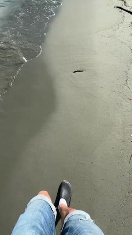 The video begins with a close-up view of a person's legs, clad in denim shorts and black slip-on shoes with white socks, as they walk along a sandy beach. The camera angle is from a first-person perspective, looking down at the legs and feet. The sand is damp and appears to be dark gray or brown in color, with subtle ripples indicating the receding tide. To the left, gentle waves lap at the shore, creating a foamy white edge where the water meets the sand. A few small pieces of driftwood are scattered on the beach. The water is a murky greenish-gray, reflecting the bright sunlight. As the person walks, their shadow is cast onto the sand. The footage captures the motion of walking, with the legs moving in rhythm and the shoes leaving faint impressions in the wet sand. The video pans slightly to the right at one point, revealing more of the beach and a bit of dry sand further up. The overall impression is that of a casual stroll along a coastline on a sunny day.
