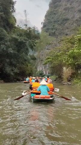 A line of small boats carrying people is being rowed along a river.  The people in the boats are wearing orange life vests and some are wearing traditional Asian hats. The boats are moving slowly through a river between lush green hills and trees. The river water appears to be murky.  The sky above is overcast.  The camera view follows one of the boats from behind.