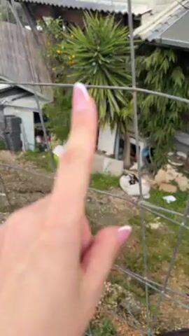 A woman's hand with manicured nails is shown reaching towards a metal fence. The camera then pans up and to the right, revealing a panoramic view of a green, hilly landscape with some buildings and a road visible in the distance. The sky is overcast with patches of blue. A bare tree is visible on the left side of the frame.