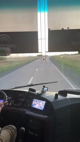 The video shows the interior of a bus from the perspective of the driver. The driver's hand is visible on the steering wheel. The dashboard is equipped with various buttons and a digital screen displaying two people. Outside the bus, a road stretches ahead into the distance, with fields on either side. The sky appears to be overcast or at dusk. The bus is moving along the road.