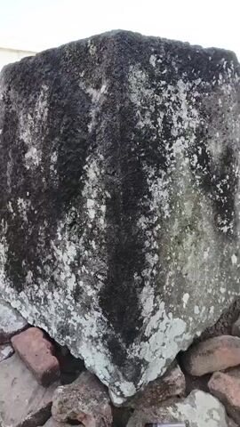 The video shows a close-up of a large, weathered stone block with dark patches and white, lichen-like growth. The camera then pans out to reveal more of these stone blocks arranged in an outdoor setting. In the background, there are some buildings with tiled roofs, a stone wall, and trees. The ground is covered with a mixture of flat stones and smaller rocks. The sky is clear and blue with some clouds. The scene appears to be an archaeological site or a historical monument. The lighting suggests it is daytime with the sun providing natural illumination. There are no people visible in the video.