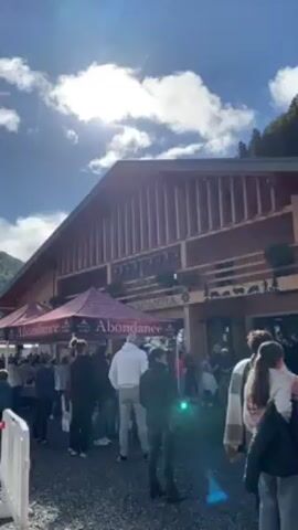 The video shows a large crowd of people gathered in front of a building with a prominent wooden facade. The building has a steeply pitched roof and vertical wooden slats creating a textured appearance. "Abondance" is written on the building. A few branded canopies with the word "Abondance" are visible on the left side of the frame. People are milling around, some standing in groups, others walking. The ground appears to be covered with gravel. The sky is partly cloudy with the sun shining through. A wooded hillside is visible in the background.