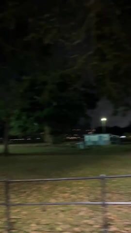 At night, the camera pans across a park. The area is grassy, with trees scattered throughout. There is a black metal fence between the viewer and the park. A red park bench is visible. Cars are parked at the far end of the park and streetlights are illuminated.