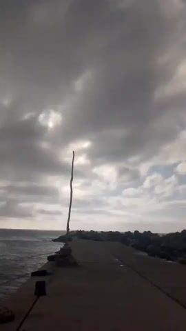 A vertical wooden pole stands in the center of a concrete pier. The pier extends into the ocean on the right side of the frame. On the left side of the pier, the ocean is visible. The sky is cloudy with patches of sunlight breaking through. Rocks line the outer edge of the pier on the right. The overall lighting is dim and overcast.