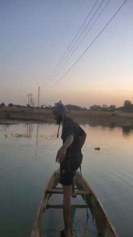 A man in a black shirt and shorts is standing in a narrow wooden boat on a calm body of water, possibly a lake or wide river. He is using a long pole to propel the boat forward by pushing off the bottom. The sky is a gradient of light blue to orange, indicating either sunrise or sunset. There are power lines overhead and some land with vegetation and buildings in the background. The water reflects the colors of the sky. The man appears to be alone in the boat.