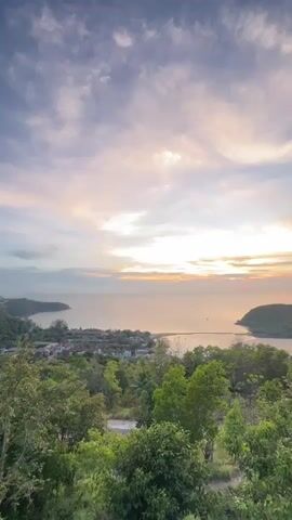 The video shows a high-angle view of a sunset scene. The sky is filled with light gray clouds, some tinged with pastel colors from the setting sun. Below the sky is a body of water, possibly the ocean, reflecting the sunlight with a golden hue. Two landmasses enclose the water, creating a bay. In the foreground is lush green vegetation, with trees obscuring the bottom of the frame. Buildings can be seen clustered along the coast. Dance music plays throughout the video.