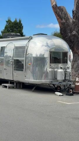 The video shows a vintage Airstream trailer parked on a paved surface. The trailer is silver and highly reflective. The front of the trailer has a curved shape with windows and propane tanks attached. A door on the side of the trailer is open, revealing steps leading into the interior. A tree trunk is visible on the right side of the frame, and a tree and blue sky with clouds are in the background. The trailer appears to be stationary.