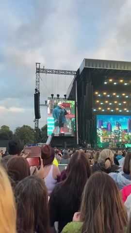 The video shows a crowd of people at an outdoor concert. A large screen displays a close-up of a man wearing a white cap and denim jacket, with green dyed hair and face paint. The stage behind him is lit with green and blue lights, and a band can be seen performing. People in the crowd are holding up their phones to record the event. The sky is overcast.