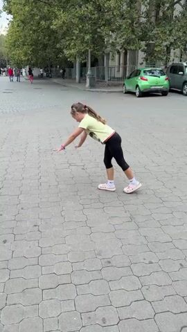 This media file captures a young girl performing a handstand and subsequent acrobatic maneuvers on a paved outdoor area. The setting appears to be a public walkway or plaza, evidenced by the patterned stone paving and the presence of trees and buildings in the background. 

The girl, wearing a yellow t-shirt, black leggings, and pink Crocs, is the sole prominent subject. She is actively engaged in gymnastic movements, starting with a handstand where her legs are extended upwards, then transitioning into a more grounded pose before standing upright. Her facial expression suggests engagement and perhaps enjoyment of her activity.

In the background, other people can be seen walking in the distance, adding to the impression of a public space. A bright green car is parked to the right of the frame, and other vehicles are partially visible. The foliage from trees suggests a warm season. The lighting implies it is daytime, possibly late morning or afternoon.

There is no visible text within the media file. The overall scene depicts a moment of spontaneous physical activity by a child in an urban or semi-urban environment.