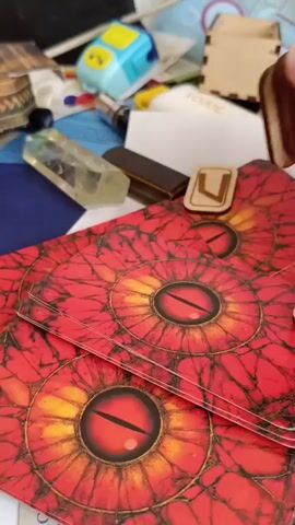 A close-up shot shows a hand placing several small, rectangular wooden runes with carved symbols onto a red surface with a pattern resembling a dragon's eye. The runes are light brown and have black symbols etched into them. In the background, various craft supplies are visible, including a clear crystal, a blue pencil sharpener, and other wooden pieces. The lighting is bright, and the focus is on the runes and the surface they are being placed on.