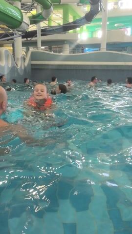 The video shows an indoor water park with a large pool filled with people. Many are wearing life vests. There are waterslides and other water features visible above the pool. The water has a light blue tint, with ripples across the surface. The water park setting is brightly lit. The camera moves slightly, capturing a wide view of the crowded pool area.