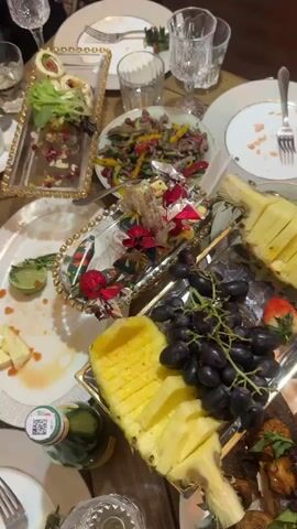 The video shows a table laden with food, suggesting a gathering or a meal setting. The table is wooden and the dishes are mostly white with gold accents. There are various food items, including sliced pineapple, grapes, sliced meat, cheese, fried spring rolls with a dipping sauce, salad, and what appears to be marinated fish. There are also bottles of mineral water, empty plates, forks, knives, and glasses. The food has been partly eaten, suggesting the meal is ongoing or has just finished. The lighting appears to be artificial, and the overall scene gives the impression of a fairly formal or special occasion.