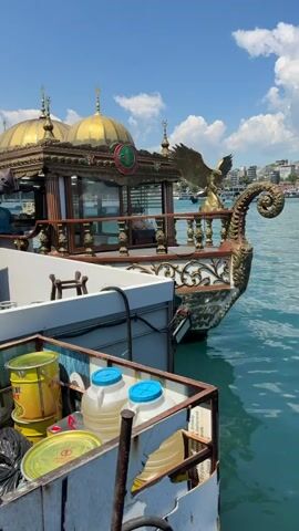 The video begins with a close-up of a boat's ornate front, featuring a golden eagle statue and a curved, decorative prow. The boat has a covered section with golden domes and intricate railings. In the foreground, on another boat or dock, are stacked yellow drums, two large plastic containers with blue lids, and a black trash bag. The water is a clear turquoise color. The camera then pans to the right, revealing a cityscape in the distance with buildings and a prominent tower, likely the Galata Tower in Istanbul. A long bridge, possibly the Galata Bridge, stretches across the water, with traffic visible on it. The sky is bright blue with scattered white clouds. There are no visible people actively engaged in any actions. The overall impression is a sunny day in a vibrant waterfront city.