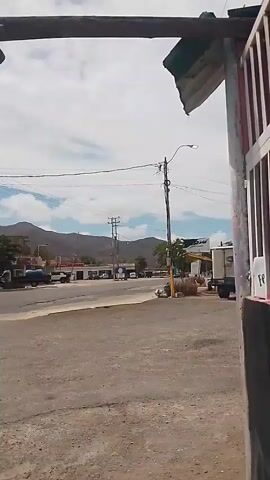 The video shows a view from under a makeshift wooden structure, looking across a gravelly area toward a road intersection. In the background, there are buildings and hills. Overhead, there are several electrical wires stretching across the scene.

A man wearing a light-colored shirt and dark shorts is walking across the road in the foreground. There are several vehicles, including cars and a motorcycle, passing through the intersection. A white vehicle is parked near a small structure with a shaded area. The sky is partly cloudy with a blue hue visible.