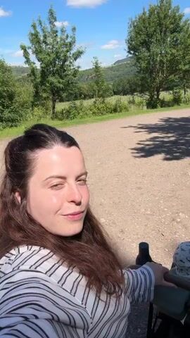 A woman in a striped shirt is in a wheelchair, smiling and looking into the camera as she is pushed by someone off-screen. The background is a sunny outdoor scene with trees and hills. The camera then turns to show the back of the woman's head as she is pushed along a gravel path.