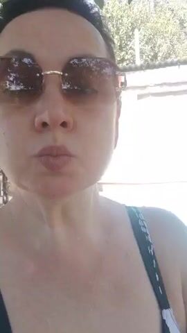 A woman wearing sunglasses and a black and white swimsuit stands in front of a swimming pool. She is looking at the camera and pouting. The pool is filled with blue water and there is a brick wall behind it. There are also trees and bushes in the background.