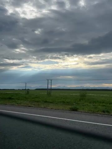 A video taken from a vehicle shows a rural landscape with a road in the foreground. A vast green field stretches out behind the road, dotted with telephone poles. In the distance, mountains form a dark silhouette against a cloudy sky. Sunlight streams through breaks in the clouds, creating visible rays of light. The video pans slowly across the scene, offering a continuous view of the field, poles, and mountains under the dramatic sky. There are no people or animals visible in the video. The environment appears to be a roadside view on a cloudy day with rays of sun breaking through.