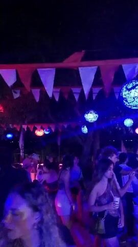 The video shows a lively outdoor party scene at night. Several people are standing and moving around, seemingly dancing or mingling near a brightly lit bar area. The bar itself is glowing with blue light. Above the crowd, there are several decorative spherical lights hanging, some glowing blue and others red.  Pennants are strung across the top of the frame. The overall atmosphere is festive and energetic.