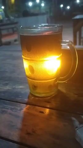The video shows a close-up of a glass mug filled with a dark liquid, possibly tea or coffee. The mug is sitting on a wooden table. A bright light source is positioned behind the mug, illuminating it from the side and creating a warm glow that passes through the liquid and the textured glass. The background is dark and appears to be outdoors at night, with blurred lights visible in the distance. There is also a hint of a cigarette pack in the lower right corner of the frame. The mug is being held up or illuminated in such a way that the light source is a focal point, casting a strong, golden light.