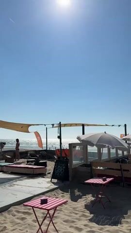 The video shows a beachside cafe on a sunny day. The camera pans from left to right, revealing several tables and seating areas on the sand. In the foreground, there are two small pink tables with chairs. Further back, there are more substantial seating areas with wooden benches and tables, some with white umbrellas providing shade. A few people are seated at these tables, appearing to be patrons of the cafe. In the middle ground, a palm tree stands in a planter. Numerous seagulls are flying in the clear blue sky above the scene, some close to the camera and others further away. The ocean is visible in the background, with a wooden barrier separating the cafe area from the beach. The overall atmosphere is relaxed and summery.