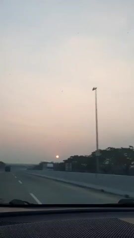 A view from inside a vehicle on a highway at sunset. The sun is visible as a small orange orb low on the horizon, partially obscured by trees. The sky is a pale, hazy color. The highway has multiple lanes separated by a concrete barrier. There are some trees and shrubbery along the side of the road. There are other vehicles in the distance. The dashboard of the vehicle is visible in the foreground.