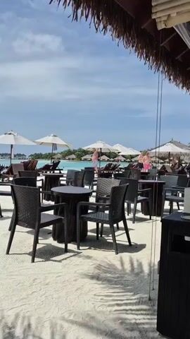 The video shows a beach scene, apparently at a resort. The sand is white, and the water is a clear turquoise. In the background, there are overwater bungalows. Several people are visible, some lounging in beach chairs under umbrellas and others walking in the water. In the foreground, there is a sandy area with dark wicker chairs and tables, seemingly part of a beach bar or restaurant. A thatched roof structure provides shade above the seating area. The sky is partly cloudy. The atmosphere is calm and peaceful.