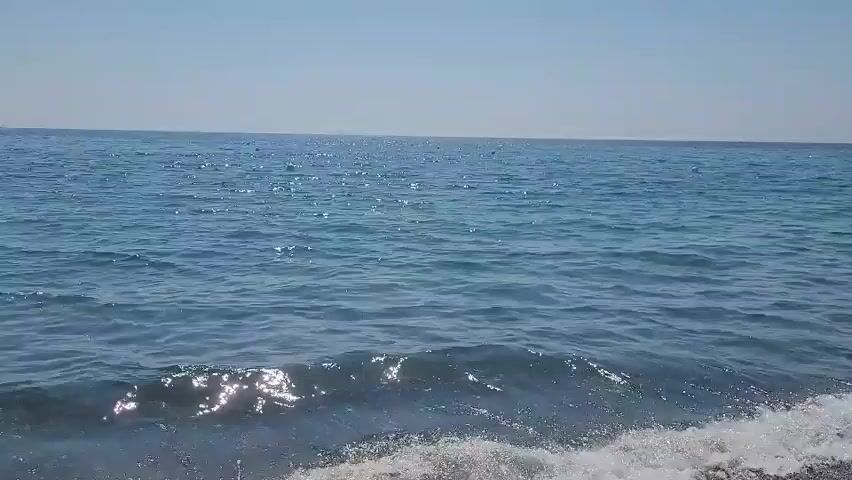 The video shows a body of water with small waves lapping at a rocky shore. The water is a deep blue and the sky is clear and bright. There are no people or objects visible in the video. The video appears to be taken on a sunny day.