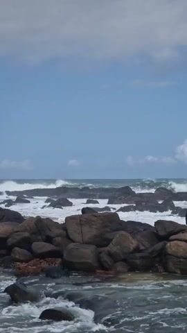 The video shows a rocky shoreline with waves crashing in the background. The foreground is composed of large, dark-colored rocks, with water flowing around them. In the distance, large waves roll in, breaking against more rocks that are scattered across the water. The sky is mostly blue with some white clouds. There are no people visible in the video. The overall scene depicts a rugged coastal environment with powerful waves.