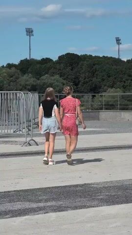 Two young women are walking away from the camera, holding hands. They are on a paved area with a dark asphalt strip. The woman on the left is wearing a black and white top and light blue denim shorts. The woman on the right is wearing a red floral dress and carrying a small beige purse. In the background, there is a line of metal fencing, a dense line of green trees, and two tall light poles. The sky is blue with scattered white clouds.