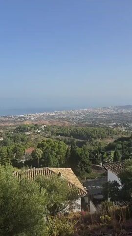 The video shows a panoramic view of a city by the sea, seen from a hillside. The foreground features the roof of a house with terracotta tiles and some green foliage. The midground consists of rolling hills covered with trees and sparse vegetation, with a road winding through it. In the background, the city sprawls along the coastline, with numerous buildings and the blue sea extending to the horizon under a clear sky. The time is daytime, indicated by the bright sunlight and the clear sky. There are no people or specific actions visible in the video. The overall scene depicts a coastal urban landscape nestled within a natural environment.