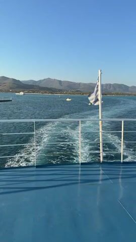 The video shows a view from the back of a boat on a sunny day with a clear blue sky. The boat is moving forward, leaving a foamy wake in the turquoise water. A Greek flag is prominently displayed on a white flagpole in the foreground, gently waving in the breeze. In the distance, a coastline with mountains can be seen, dotted with buildings and a pier. Another boat appears to be on the water in the mid-ground. The deck of the boat in the foreground is painted blue.